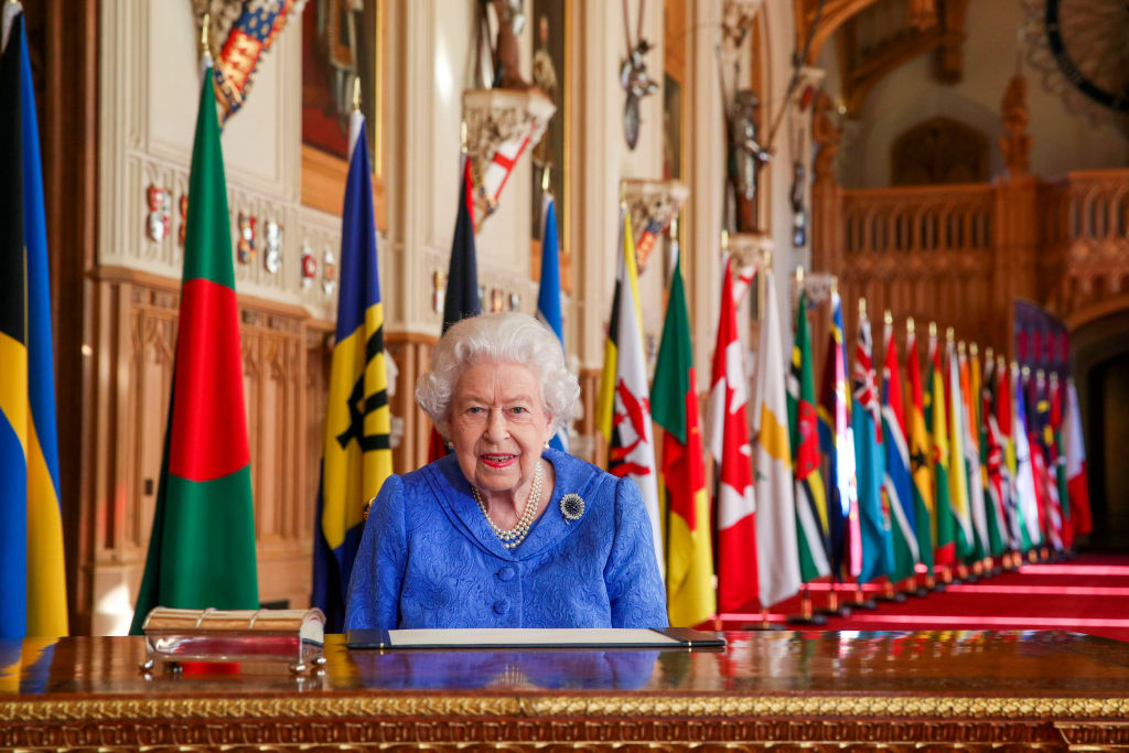 Her Majesty Queen Elizabeth II wearing a blue suit and pearls, seated at an ornate table. Behind her is a row of flags from countries of the Commonwealth.