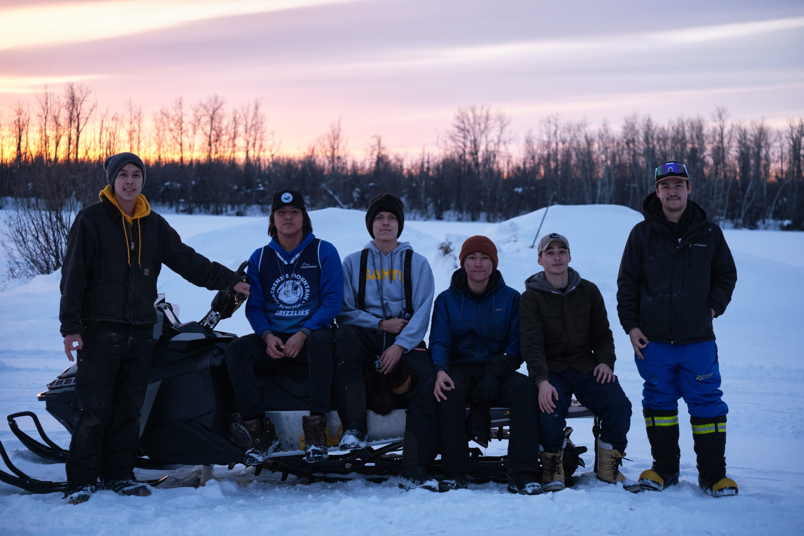 Six young people in a row, some standing, some sitting on a snow machine, in a snowy field with the sun setting in the background.
