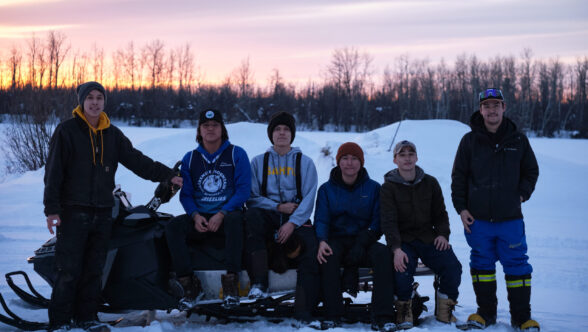 Six young people in a row, some standing, some sitting on a snow machine, in a snowy field with the sun setting in the background.