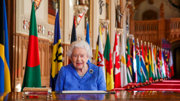 Sa Majesté la reine Elizabeth II, portant un costume bleu et des perles, assise à une table ornée. Derrière elle se trouve une rangée de drapeaux des pays du Commonwealth.