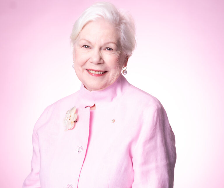 A white-haired woman wears a pale pink suit, standing in front of a pale pink background.