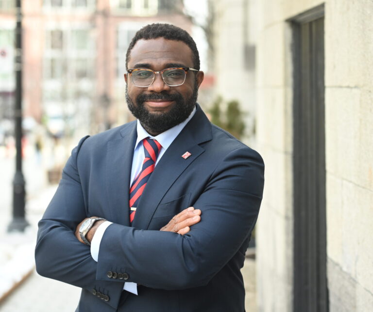 A smiling man stands in a dark blue suit and a red and blue striped tie with his arms folded across his chest.