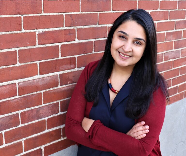 A woman with long dark hair dressed in a red sweater leans against a brick wall.