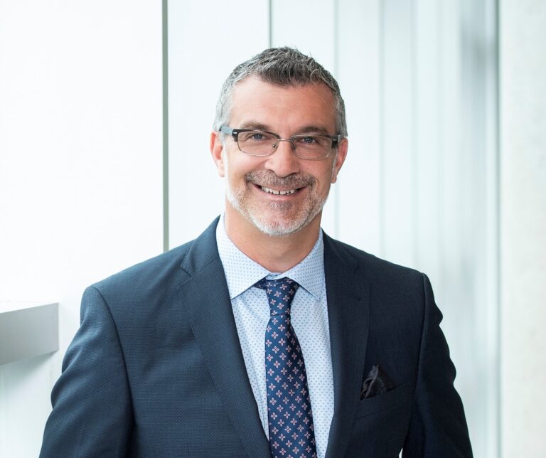 A man in a blue suit and tie with glasses smiles at the camera.