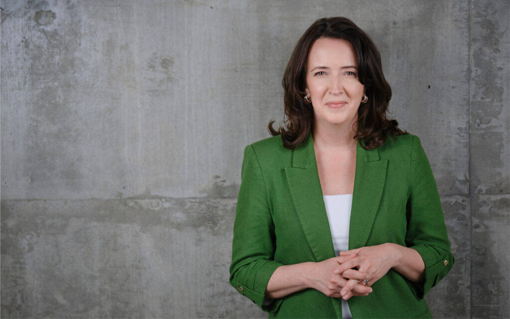 Une femme aux cheveux bruns mi-longs porte un blazer vert, les mains jointes devant elle.